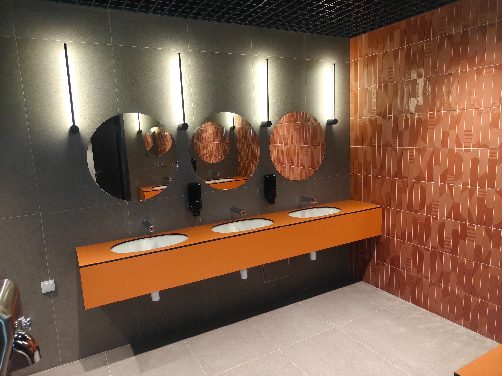 Nowoczesna łazienka publiczna z trzema okrągłymi lustrami, pomarańczową umywalką i geometrycznymi, brązowymi płytkami na ścianie. Minimalistyczne oświetlenie.