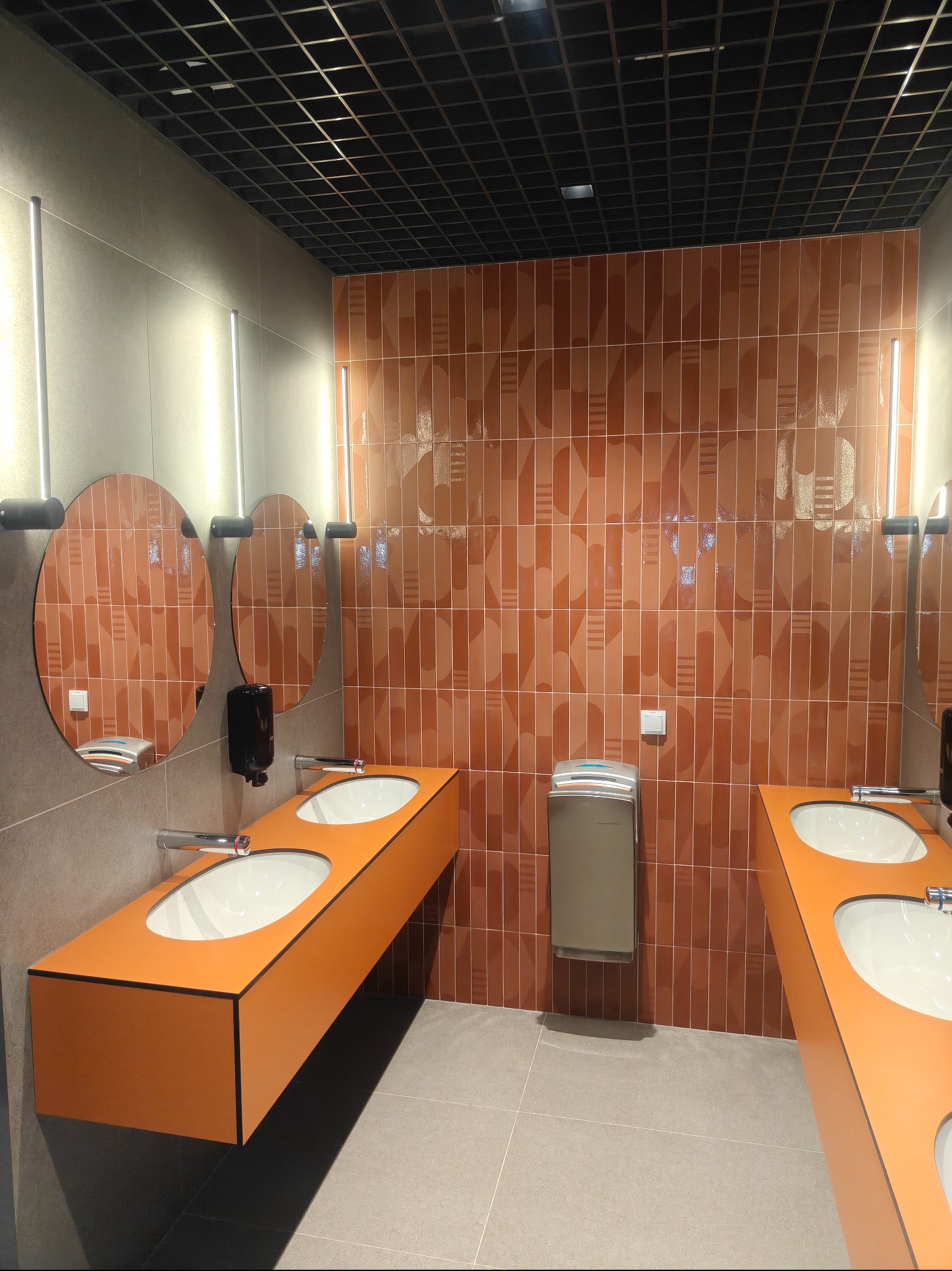 Nowoczesna łazienka publiczna z pomarańczowymi akcentami: umywalki, lustra i płytki ścienne. Minimalistyczny design, oświetlenie LED i geometryczny wzór płytek.