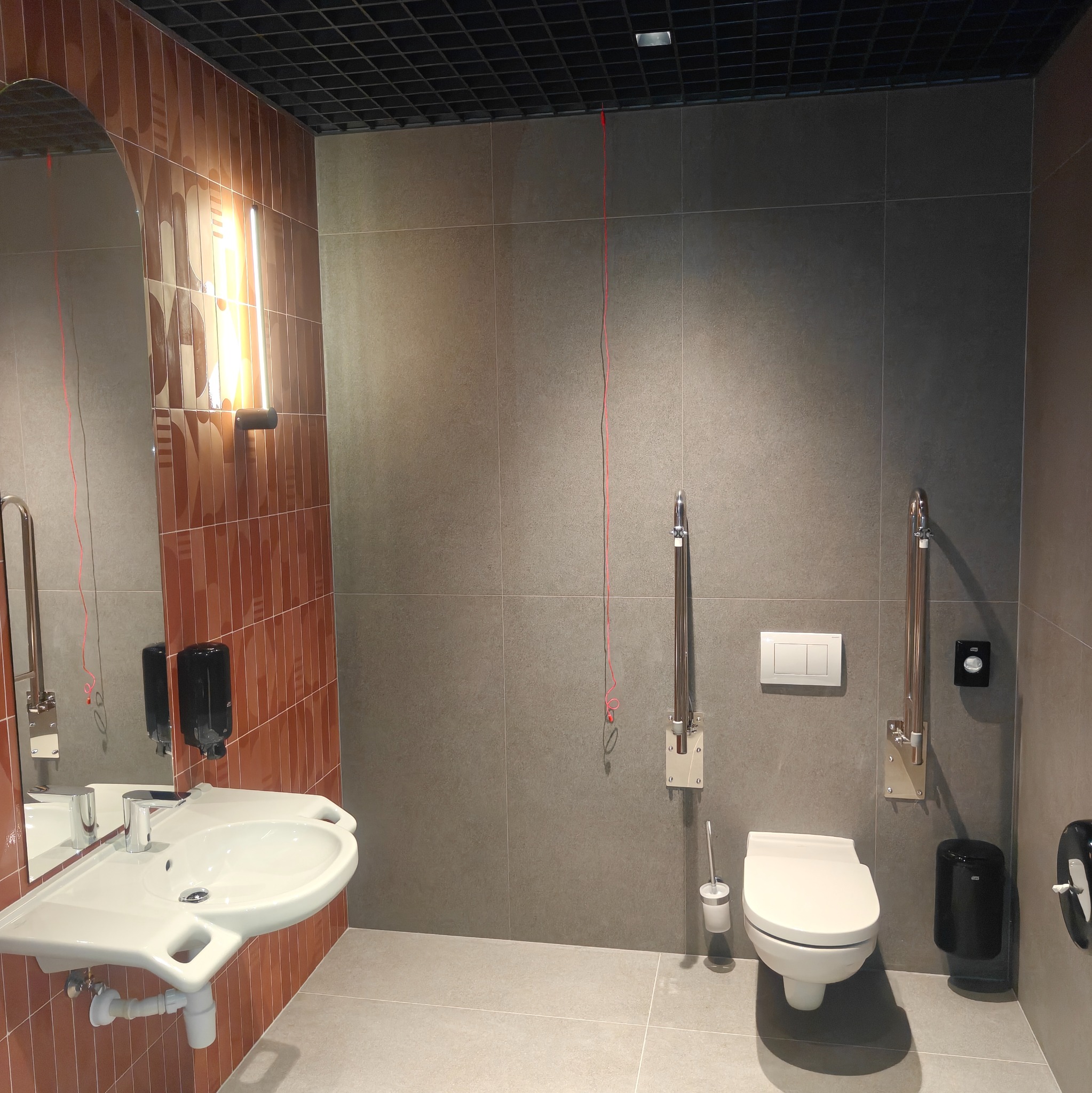 Wykończona łazienka z umywalką, toaletą i uchwytami dla niepełnosprawnych. Brązowe i szare płytki, oświetlenie LED. Widoczne akcesoria łazienkowe i lustro.