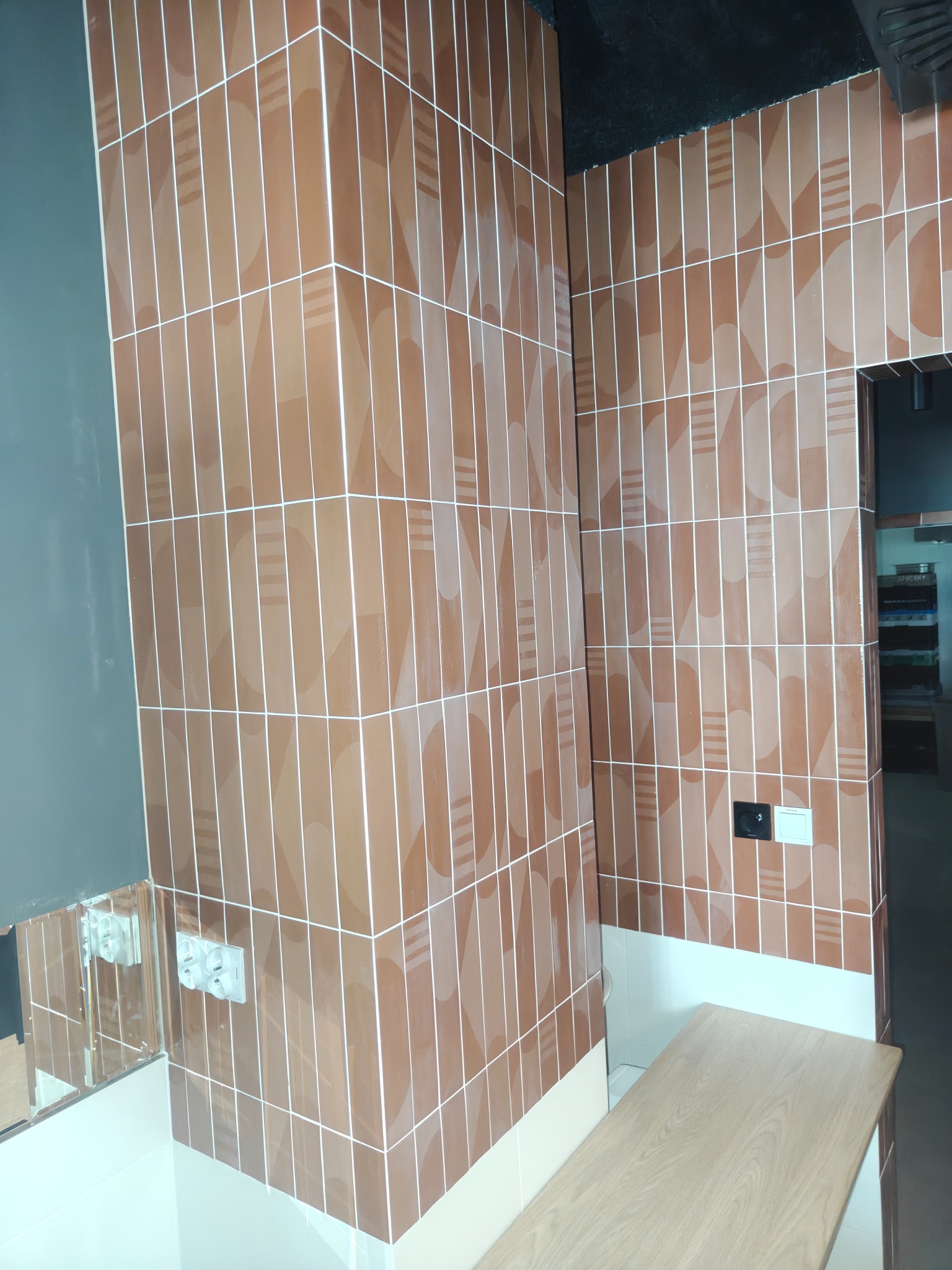 Brązowe, pionowe płytki z geometrycznym wzorem na ścianie, biała fuga, gniazdka elektryczne, fragment lustra i drewniany blat w pomieszczeniu po remoncie.