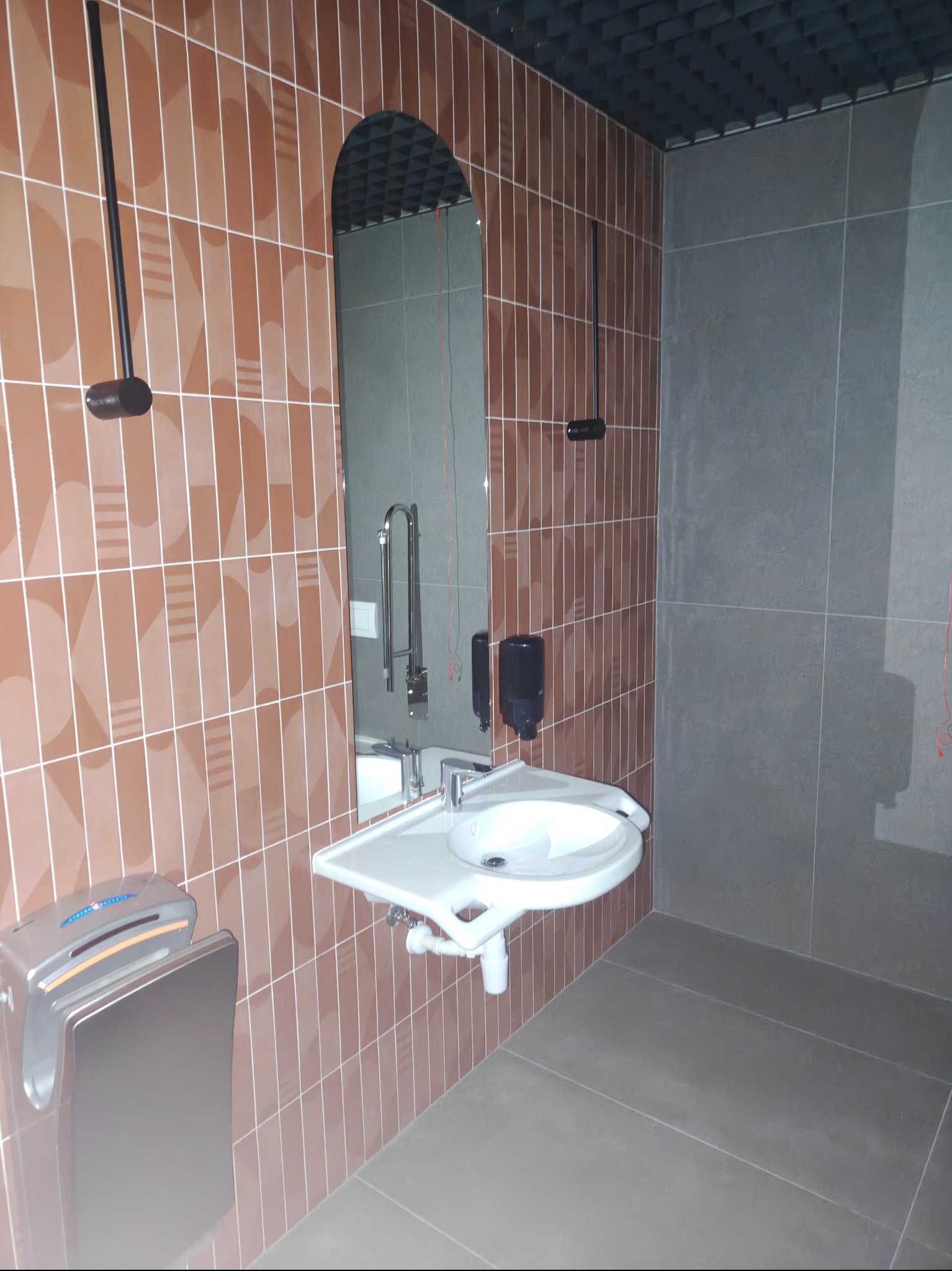 Wykończona łazienka publiczna z brązowymi płytkami, umywalką dla niepełnosprawnych, lustrem i szarą podłogą. Widoczny dozownik mydła i suszarka do rąk.