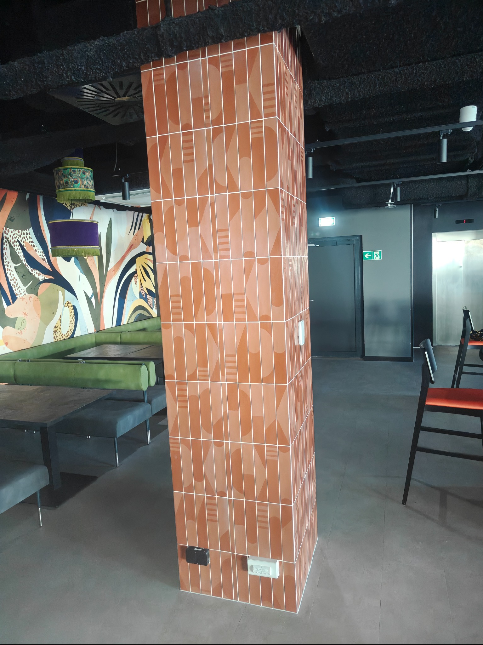 Obłożona terakotą kolumna w lokalu gastronomicznym z muralem w tle. Widoczne stoliki i krzesła, sufit z instalacjami. Nietypowy wzór płytek i oświetlenie nadają wnętrzu charakter.