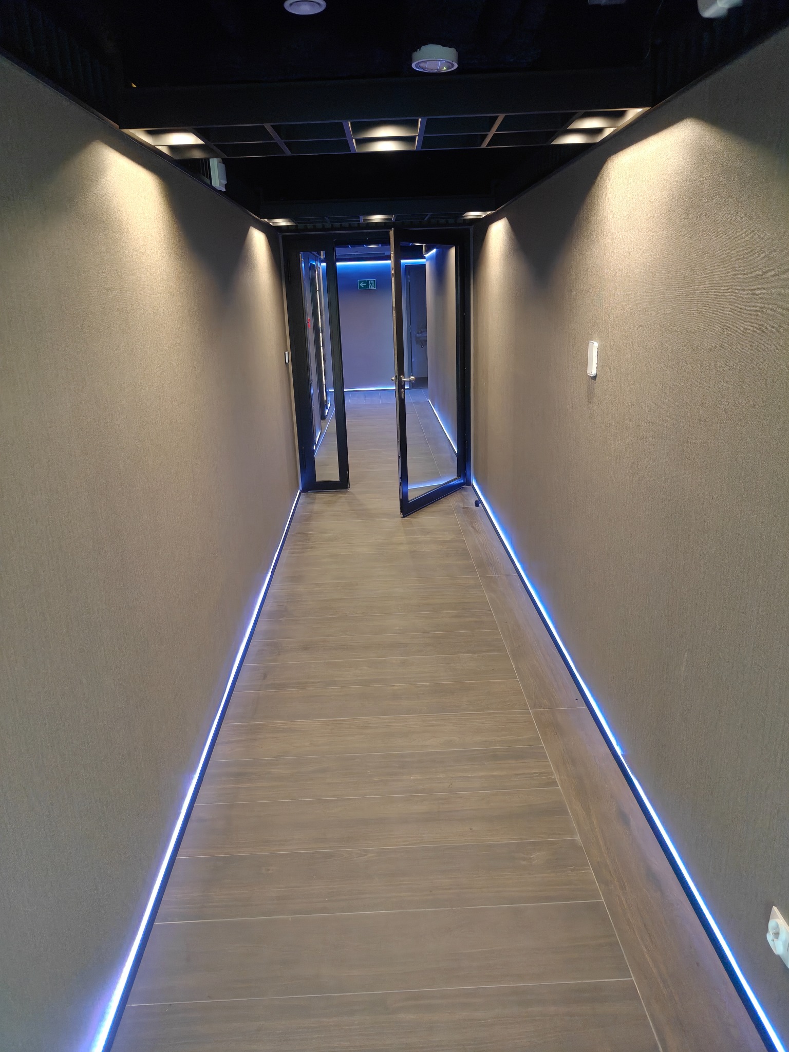 Nowoczesny korytarz biurowy z podświetleniem LED wzdłuż ścian i podłogi z paneli. Otwarta szklana drzwi na końcu korytarza. Oświetlenie sufitowe punktowe.