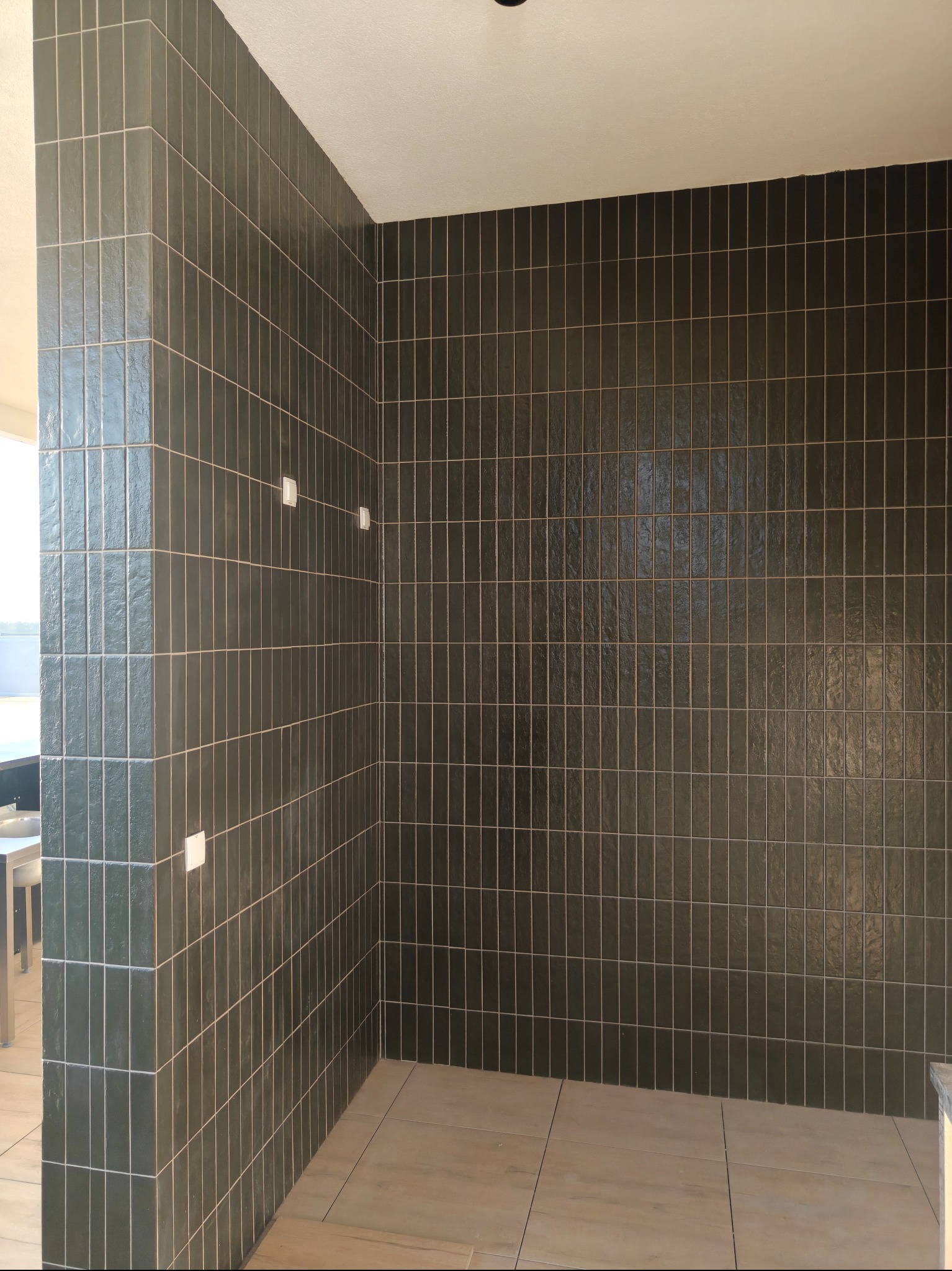 Wnętrze łazienki po remoncie: ściany wyłożone pionowymi, ciemnozielonymi płytkami, jasna podłoga. Widoczne białe elementy instalacji elektrycznej na ścianie.