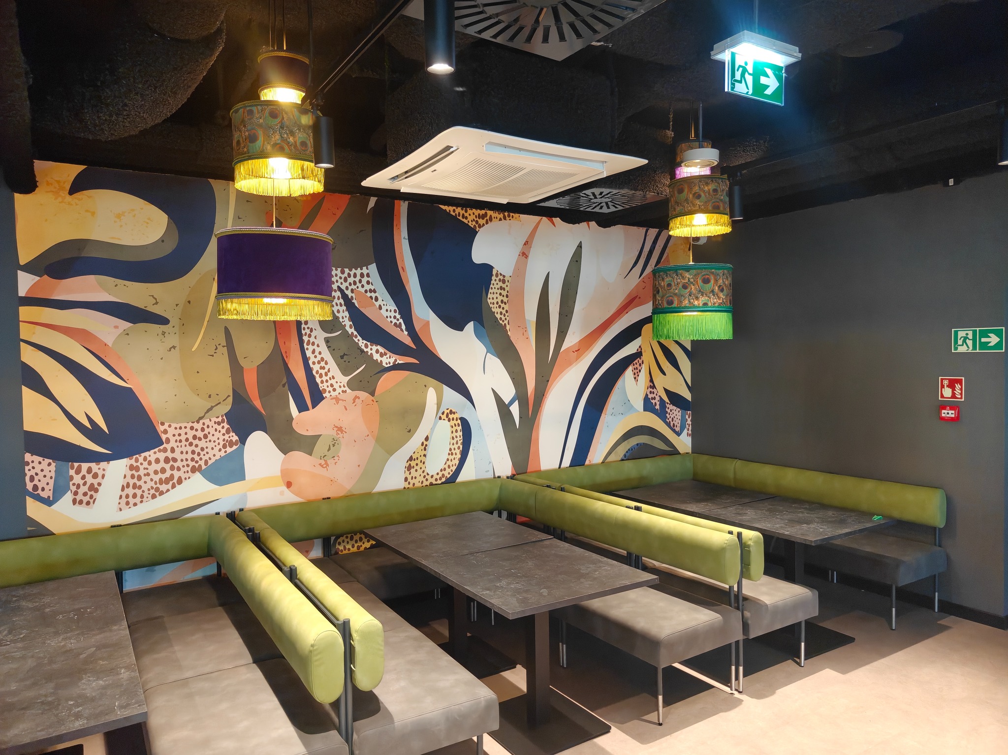 Nowoczesne wnętrze biurowe z abstrakcyjną grafiką na ścianie, ciemnymi stołami i zielonymi sofami. Dekoracyjne lampy wiszące dodają koloru i charakteru.