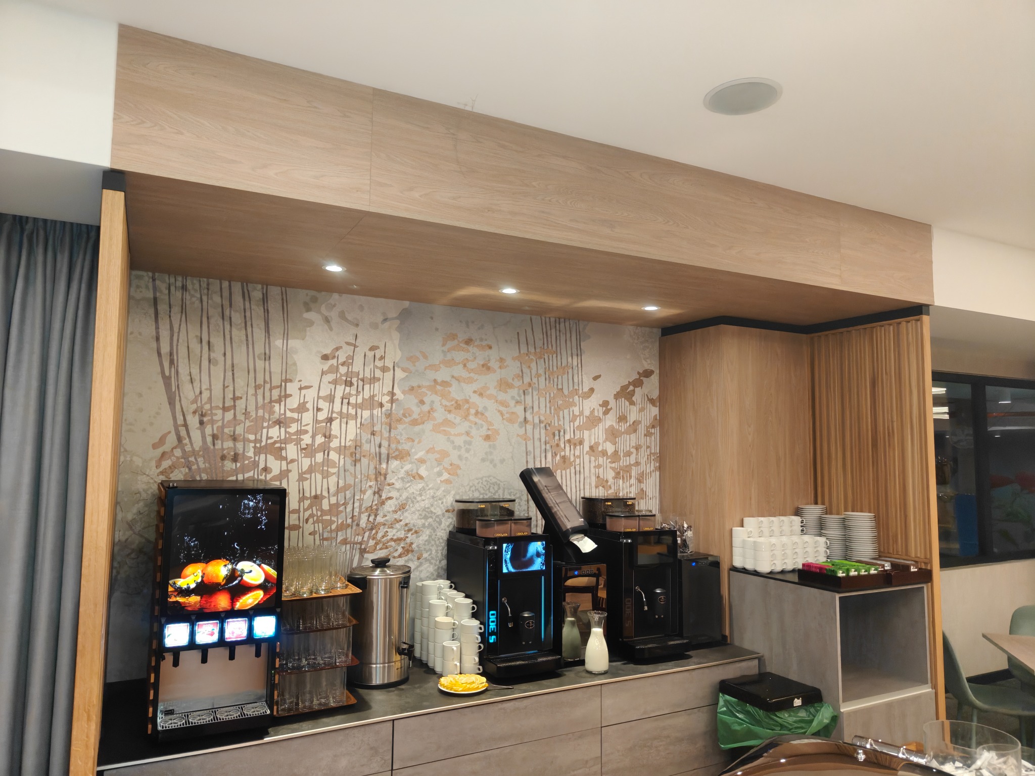 Nowoczesna strefa bufetowa z dystrybutorem napojów, ekspresami do kawy i zastawą stołową na tle ściany z motywem drzew, oświetlona punktowymi lampami.