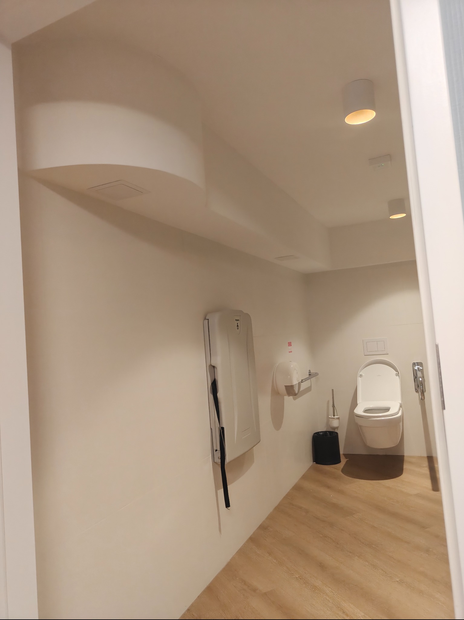 Jasne wnętrze toalety publicznej z drewnopodobną podłogą, przewijakiem dla niemowląt i minimalistycznym oświetleniem punktowym.