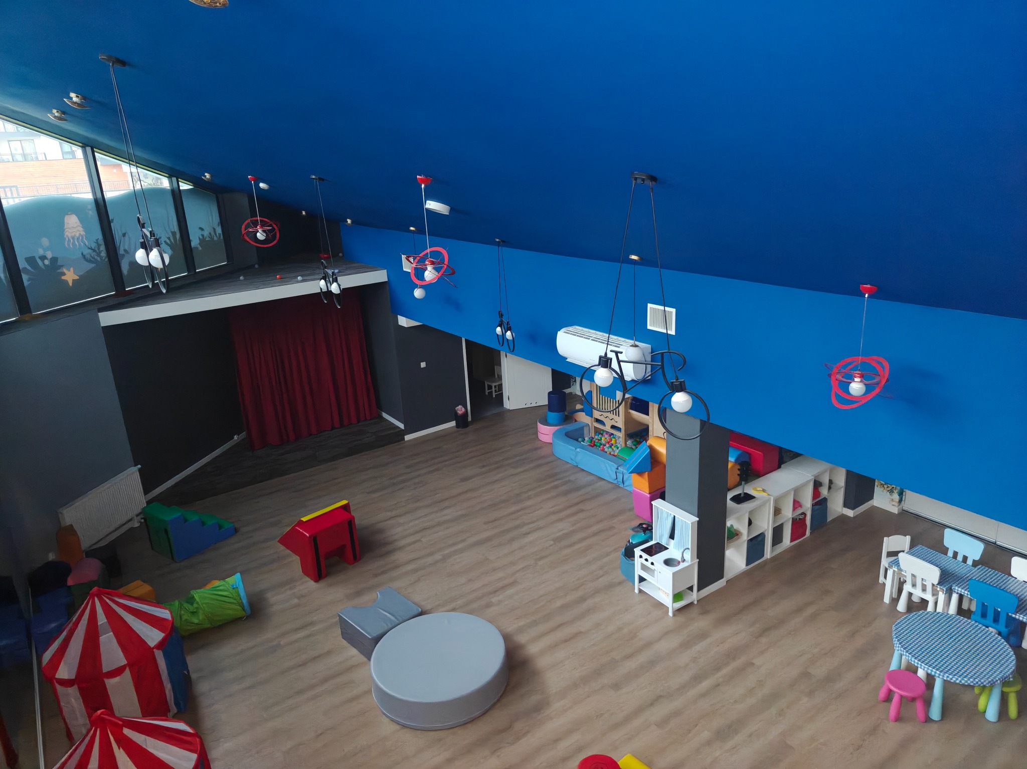 Wnętrze sali zabaw dla dzieci z niebieskim sufitem, drewnianą podłogą, małymi stolikami i krzesełkami. Widok z góry ukazuje scenę z czerwoną kurtyną.