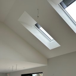 Wall Done Szymon Andrysiak - Wnętrze poddasza z dwoma oknami dachowymi z roletami, świeżo pomalowane ściany i sufit na biało, widoczne wystające przewody elektryczne.