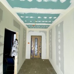 Wall Done Szymon Andrysiak - Korytarz w trakcie prac przygotowawczych do malowania w Zduńskiej Woli: ściany i sufit z widocznymi łatami szpachli, zabezpieczone drzwi wejściowe folią, widoczne przewody elektryczne na suficie...