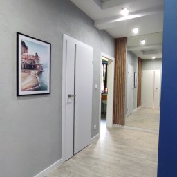 Wall Done Szymon Andrysiak - Korytarz w odcieniach szarości z panelami podłogowymi imitującymi drewno, białymi drzwiami z pionowymi wstawkami, obrazem na ścianie i dekoracyjnym sufitem podwieszanym z oświetleniem punktowym.