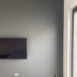 Wall Done Szymon Andrysiak - Ściana w odcieniach szarości z zamontowanym telewizorem, widoczne gniazdka elektryczne i otwór na kabel, drewniana podłoga oraz fragment okna z białymi ramami.