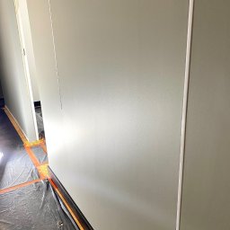 Wall Done Szymon Andrysiak - Świeżo pomalowana ściana w odcieniach szarości z widocznymi listwami przypodłogowymi, zabezpieczona folią malarską na podłodze, w trakcie prac wykończeniowych.