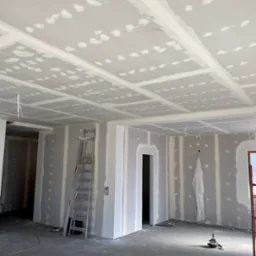 Wnętrze w trakcie prac wykończeniowych: ściany i sufit pokryte płytami gipsowo-kartonowymi ze wstępnie nałożoną warstwą szpachli, widoczna drabina aluminiowa i narzędzia na podłodze.