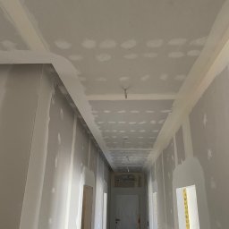 Wall Done Szymon Andrysiak - Korytarz w trakcie prac wykończeniowych, ściany i sufit pokryte warstwą szpachli z widocznymi punktowymi poprawkami, przygotowane do dalszego etapu.