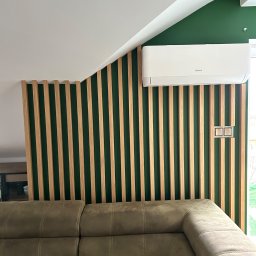 Wall Done Szymon Andrysiak - Nowoczesne wnętrze z zieloną ścianą, drewnianymi lamelami, klimatyzatorem marki Mistral i beżową sofą.