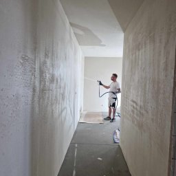 Wall Done Szymon Andrysiak - Aplikacja gładzi natryskowej na ścianę w korytarzu, widoczny pracownik z pistoletem natryskowym i ślady świeżej gładzi.