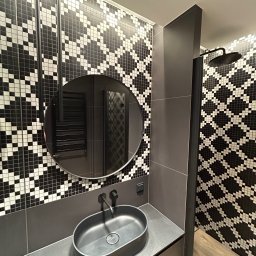 Wall Done Szymon Andrysiak - Nowoczesna łazienka w odcieniach szarości i czerni z mozaiką na ścianach, owalną umywalką na blacie i okrągłym lustrem, widoczny fragment kabiny prysznicowej z deszczownicą.