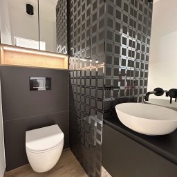 Wall Done Szymon Andrysiak - Nowoczesna łazienka z czarnymi, wzorzystymi płytkami na ścianie, białą umywalką nablatową z czarną armaturą, podwieszaną toaletą i drewnopodobną podłogą.