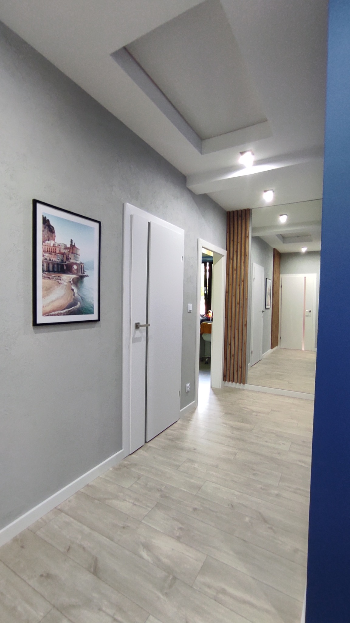 Korytarz w odcieniach szarości z panelami podłogowymi imitującymi drewno, białymi drzwiami z pionowymi wstawkami, obrazem na ścianie i dekoracyjnym sufitem podwieszanym z oświetleniem punktowym.