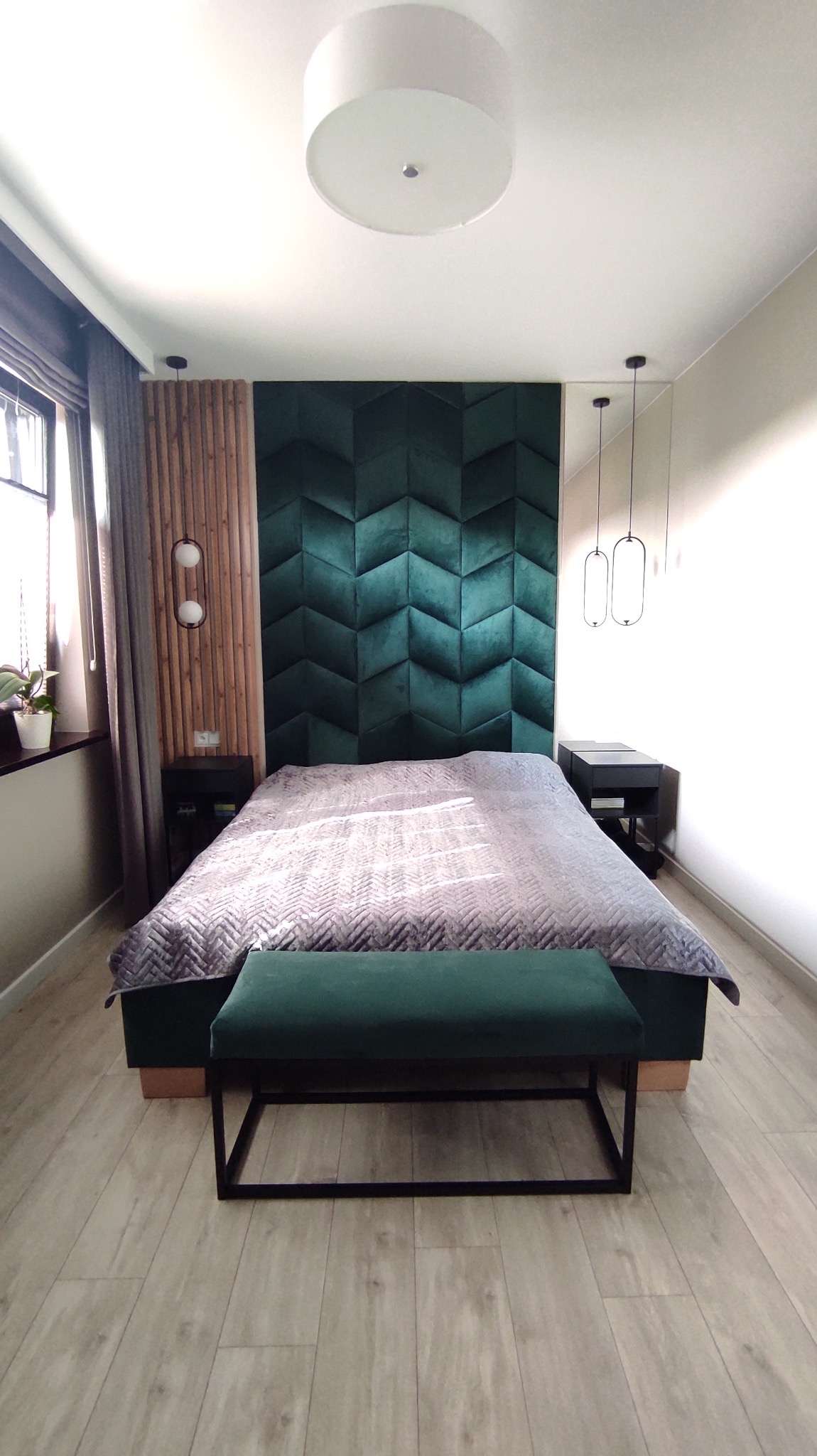 Nowoczesna sypialnia z łóżkiem, zielonym panelem ściennym w geometryczny wzór, drewnianą ścianką działową i wiszącymi lampami, wykończona w jasnych barwach.