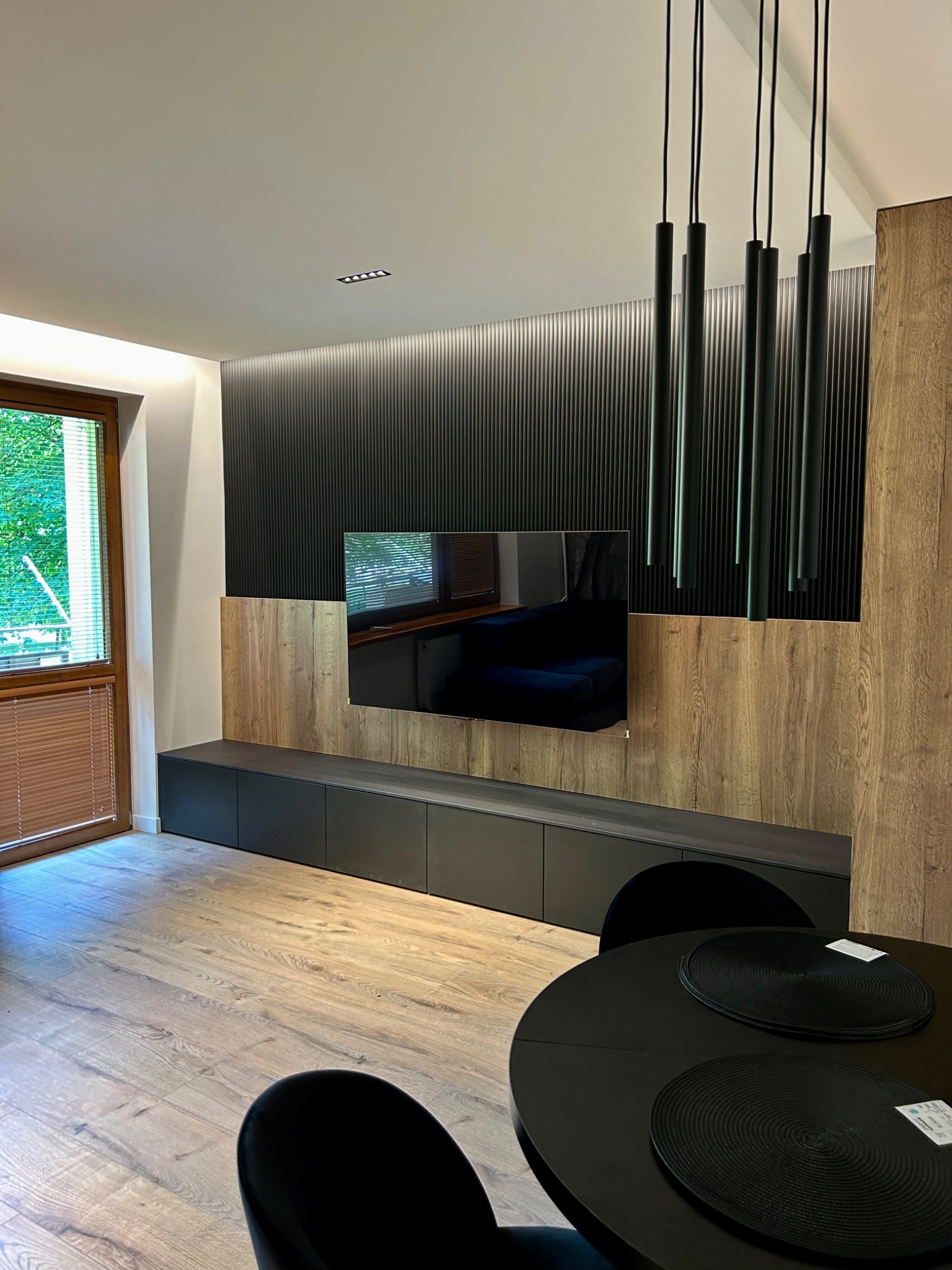 Nowoczesne wnętrze salonu z drewnianą podłogą, ścianą z pionowymi panelami, wiszącą szafką RTV i okrągłym stołem z czarnymi podkładkami.