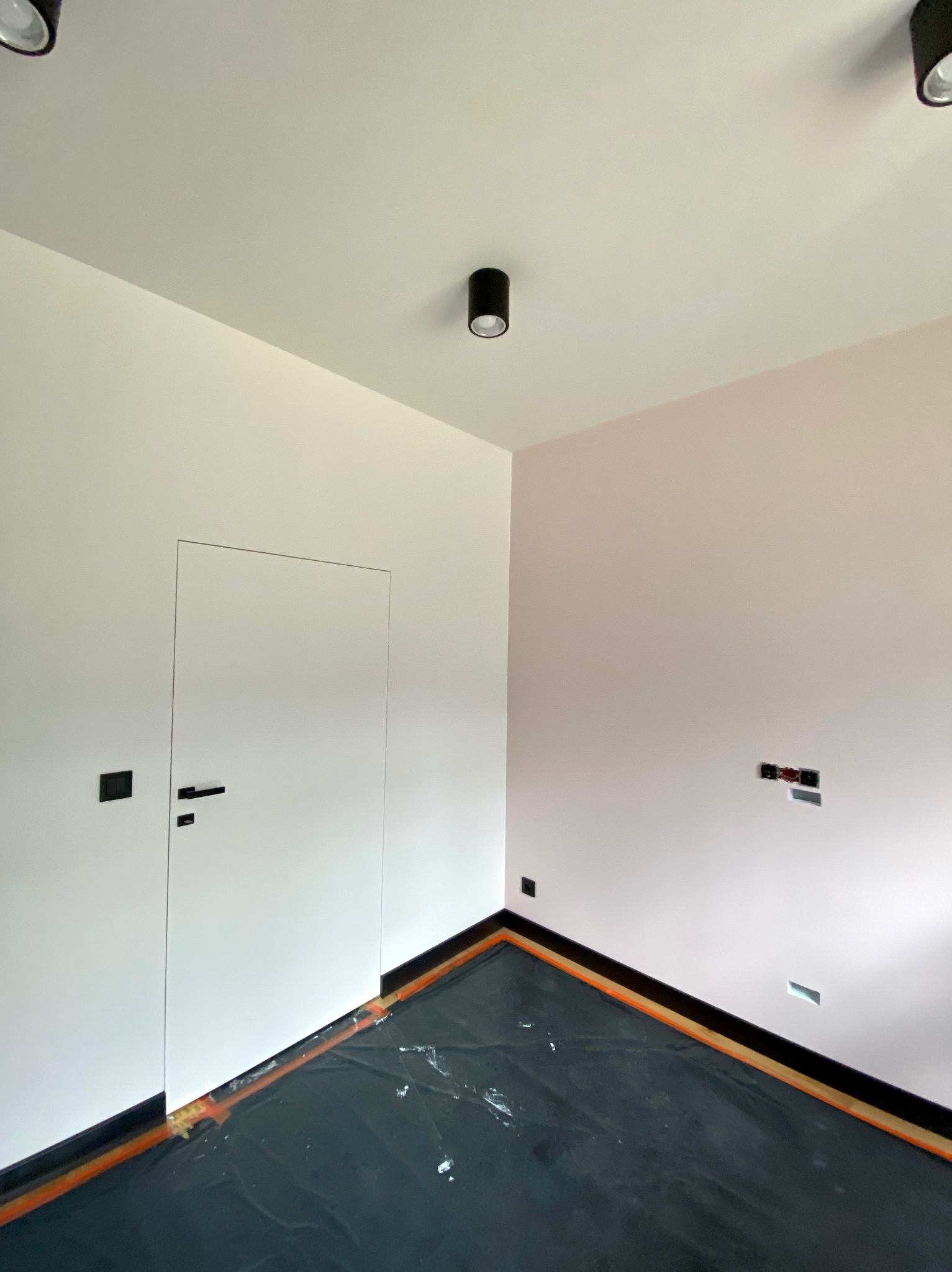 Pokój w trakcie prac wykończeniowych: ściany pomalowane na jasny róż, zabezpieczona folią podłoga z widocznymi listwami przypodłogowymi, minimalistyczne drzwi wewnętrzne i czarne lampy sufitowe.