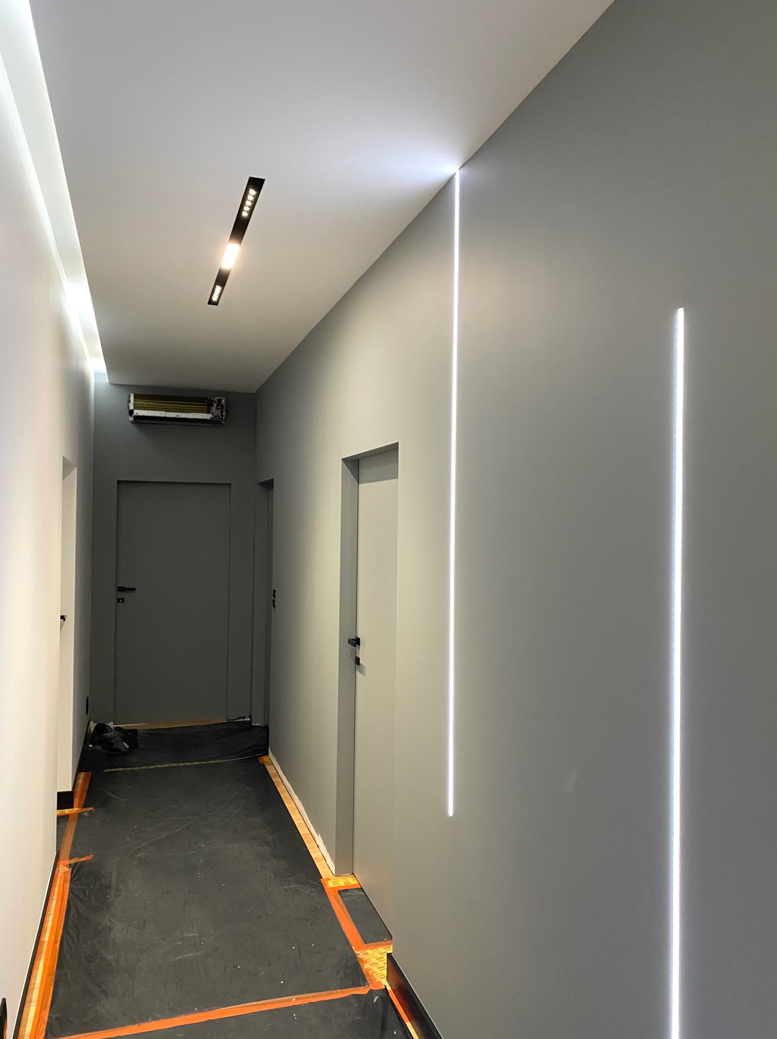Szary korytarz z zamontowanymi pionowymi liniami świetlnymi LED na ścianach, zabezpieczona podłoga folią ochronną i widoczne drzwi.