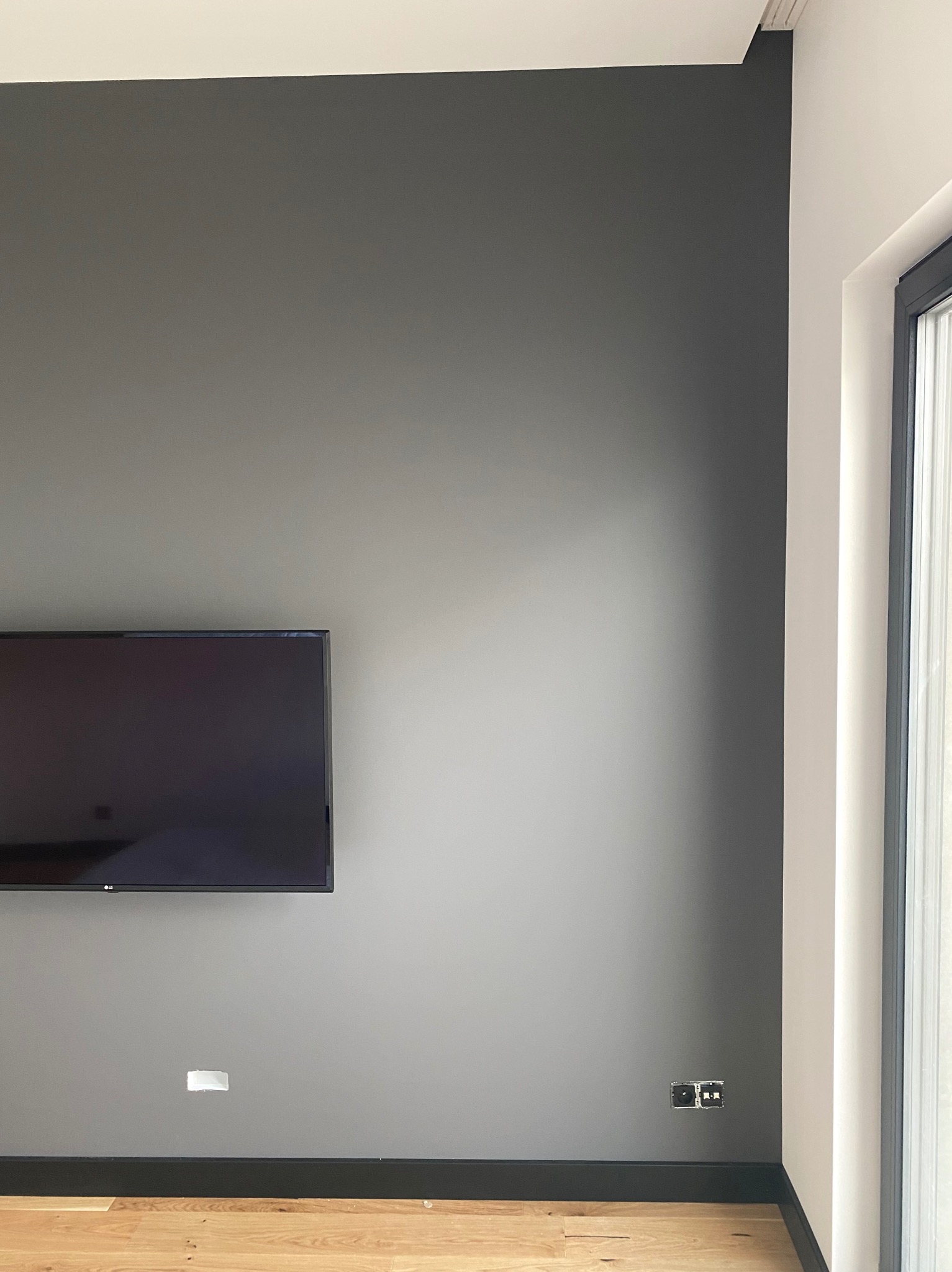Ściana w odcieniach szarości z zamontowanym telewizorem, widoczne gniazdka elektryczne i otwór na kabel, drewniana podłoga oraz fragment okna z białymi ramami.