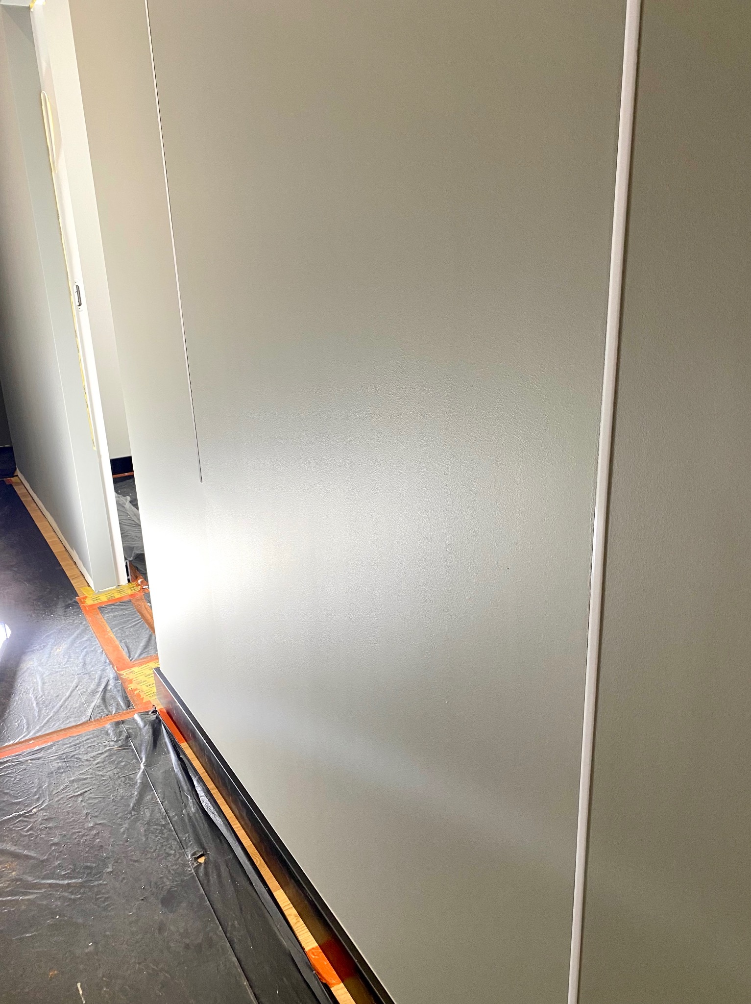 Świeżo pomalowana ściana w odcieniach szarości z widocznymi listwami przypodłogowymi, zabezpieczona folią malarską na podłodze, w trakcie prac wykończeniowych.