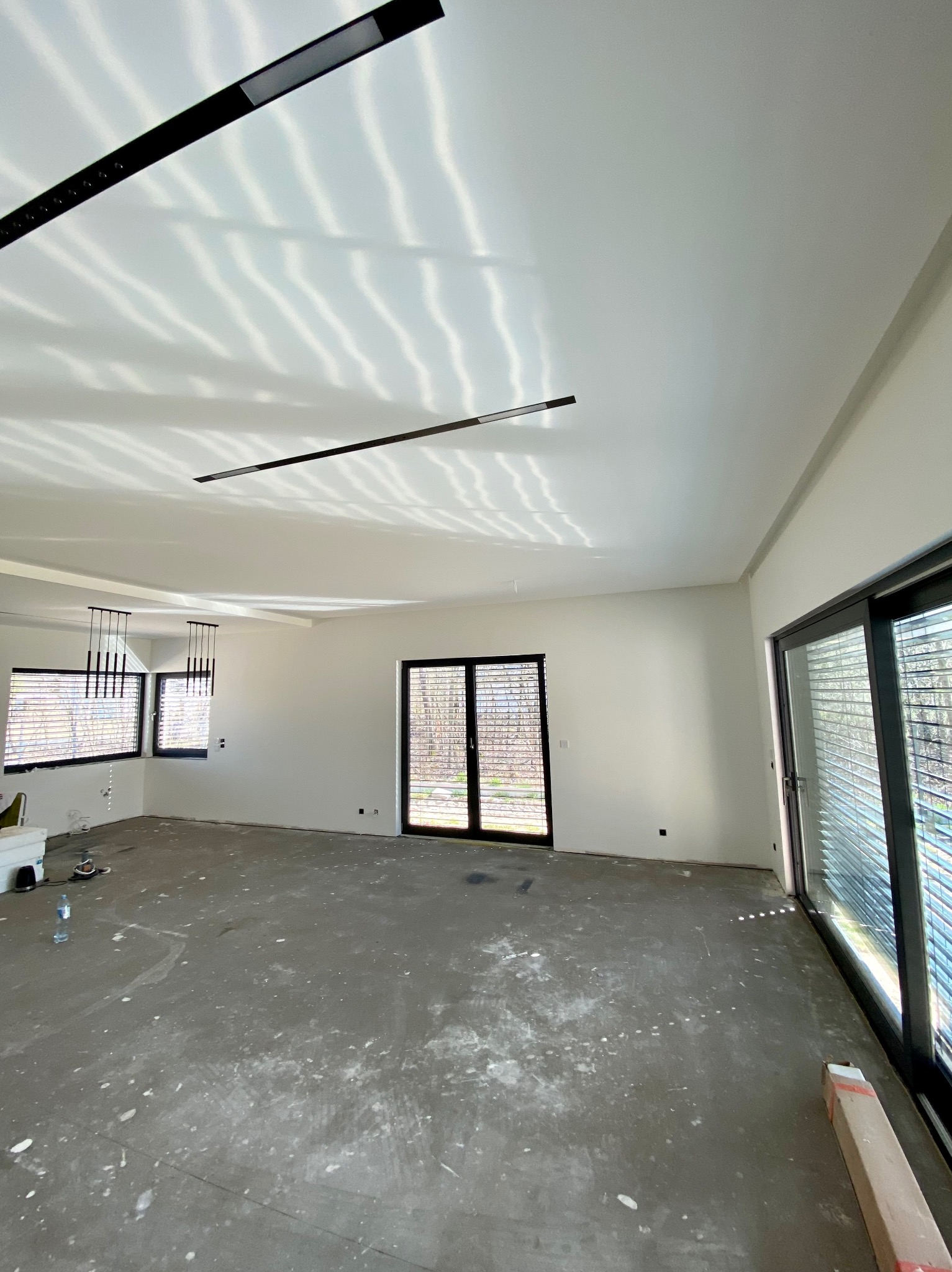 Wnętrze w stanie deweloperskim z gładzią na ścianach i suficie, widoczne ślady po pracach wykończeniowych na podłodze, nowoczesne oświetlenie sufitowe, duże okna z żaluzjami.