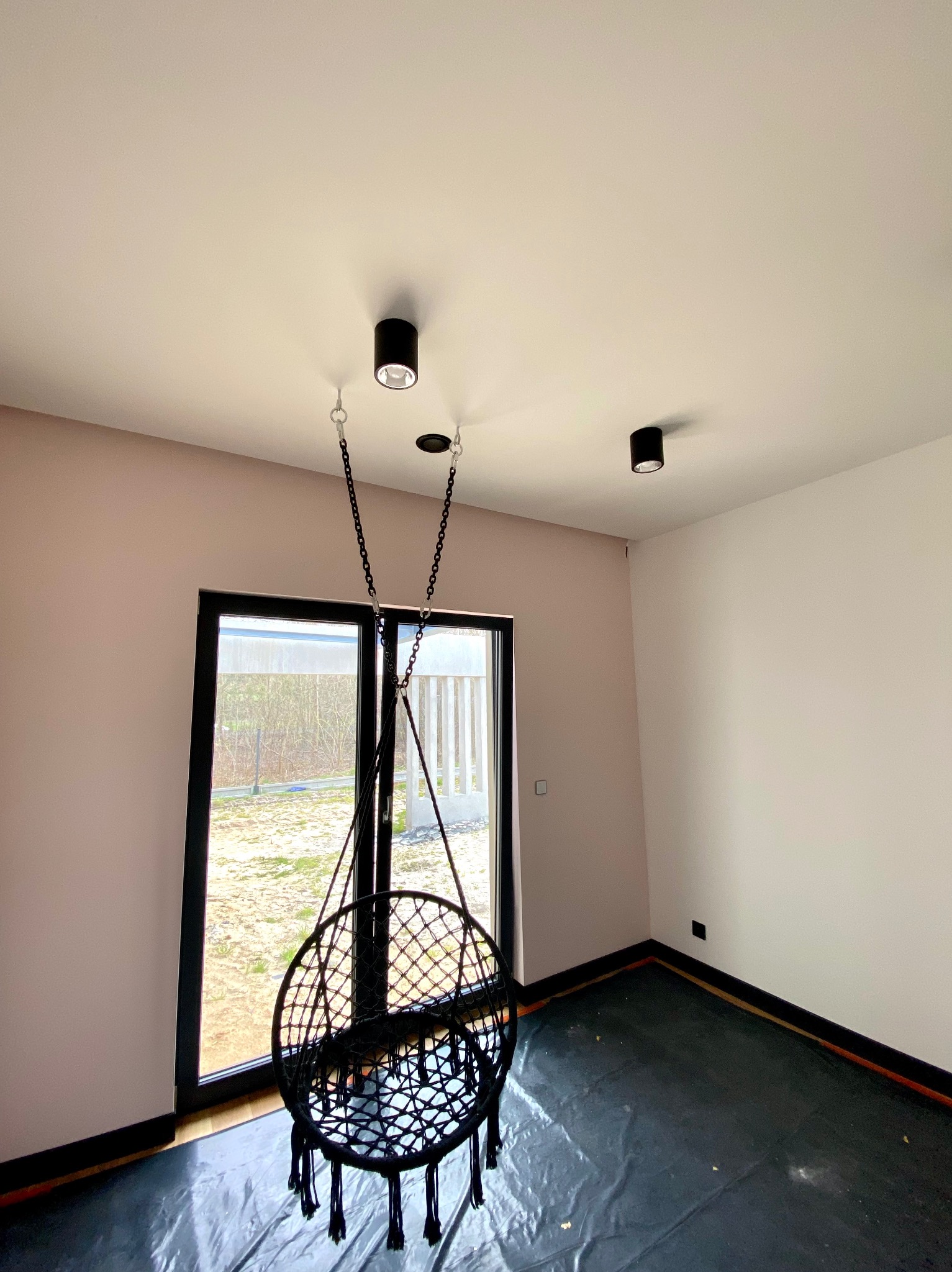 Pokój po malowaniu ścian na dwa kolory, z zamontowanym fotelem wiszącym na łańcuchu i lampami sufitowymi, podłoga zabezpieczona folią malarską.