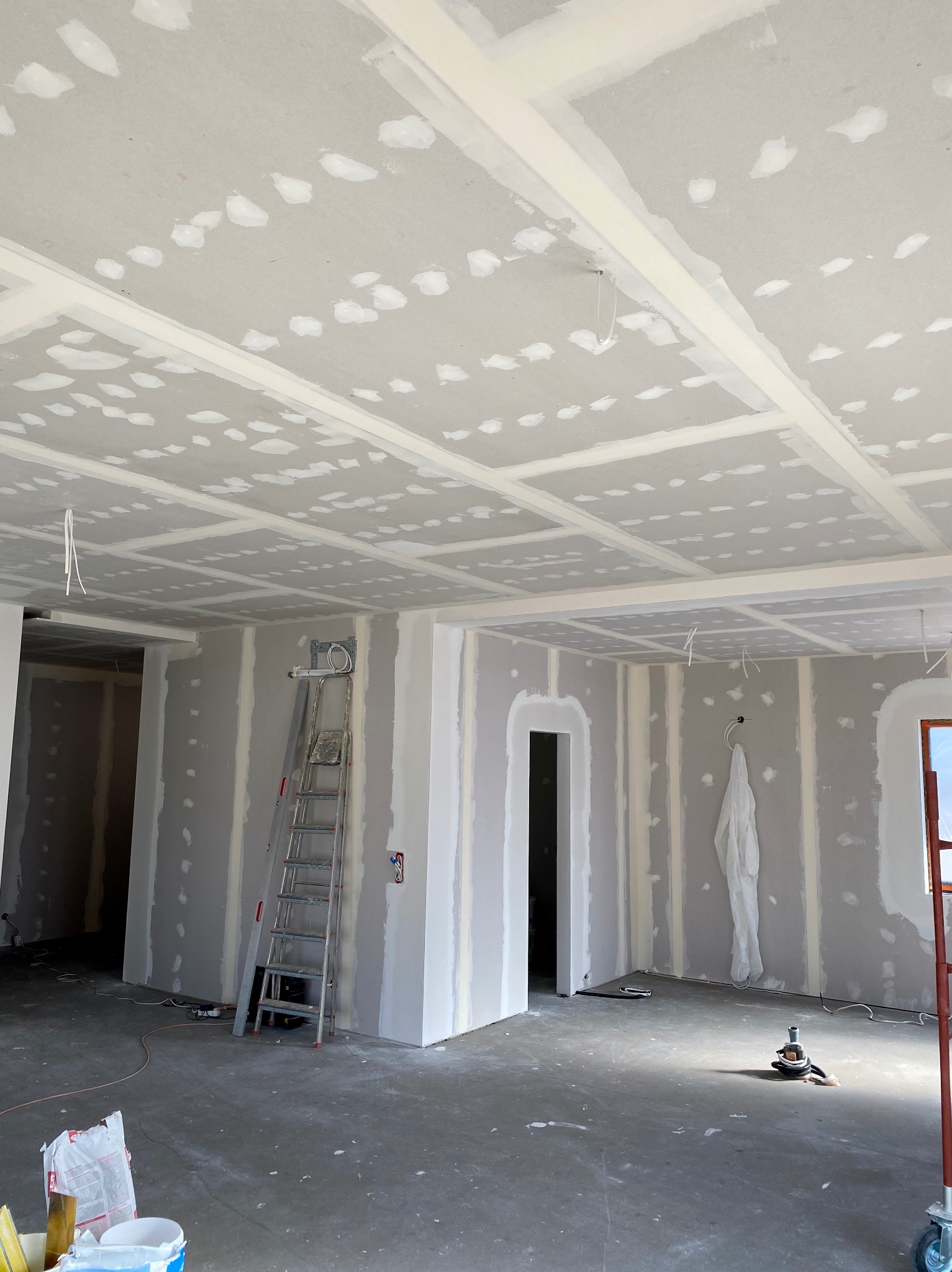 Wnętrze w trakcie prac wykończeniowych: ściany i sufit pokryte płytami gipsowo-kartonowymi ze wstępnie nałożoną warstwą szpachli, widoczna drabina aluminiowa i narzędzia na podłodze.