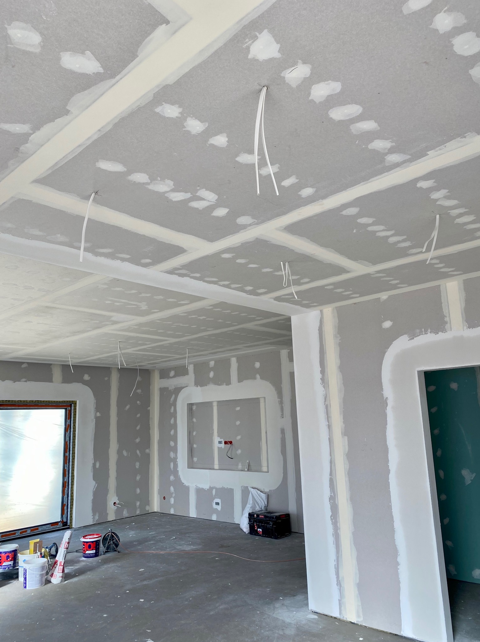 Wnętrze w trakcie prac wykończeniowych: sufit z płyt kartonowo-gipsowych z widocznymi przewodami elektrycznymi, ściany przygotowane do dalszych prac, narzędzia malarskie i wiaderka z farbą...