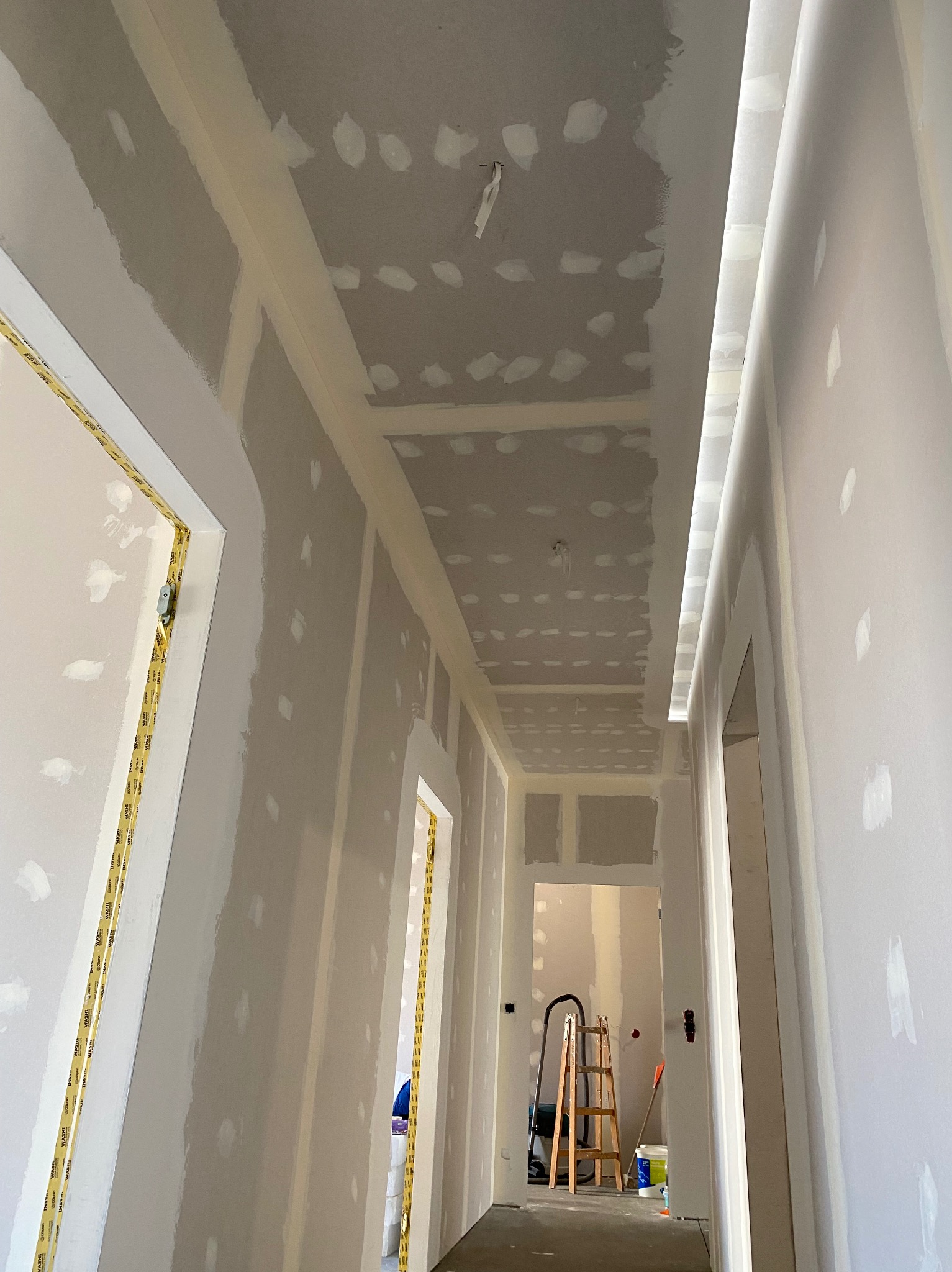 Korytarz w trakcie remontu: ściany z płyt karton-gips, widoczne punkty szpachlowania na suficie, zabezpieczone taśmą malarską ościeżnice, drabina w tle.