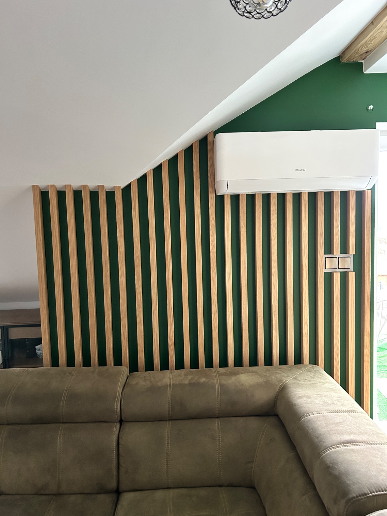 Nowoczesne wnętrze z zieloną ścianą, drewnianymi lamelami, klimatyzatorem marki Mistral i beżową sofą.