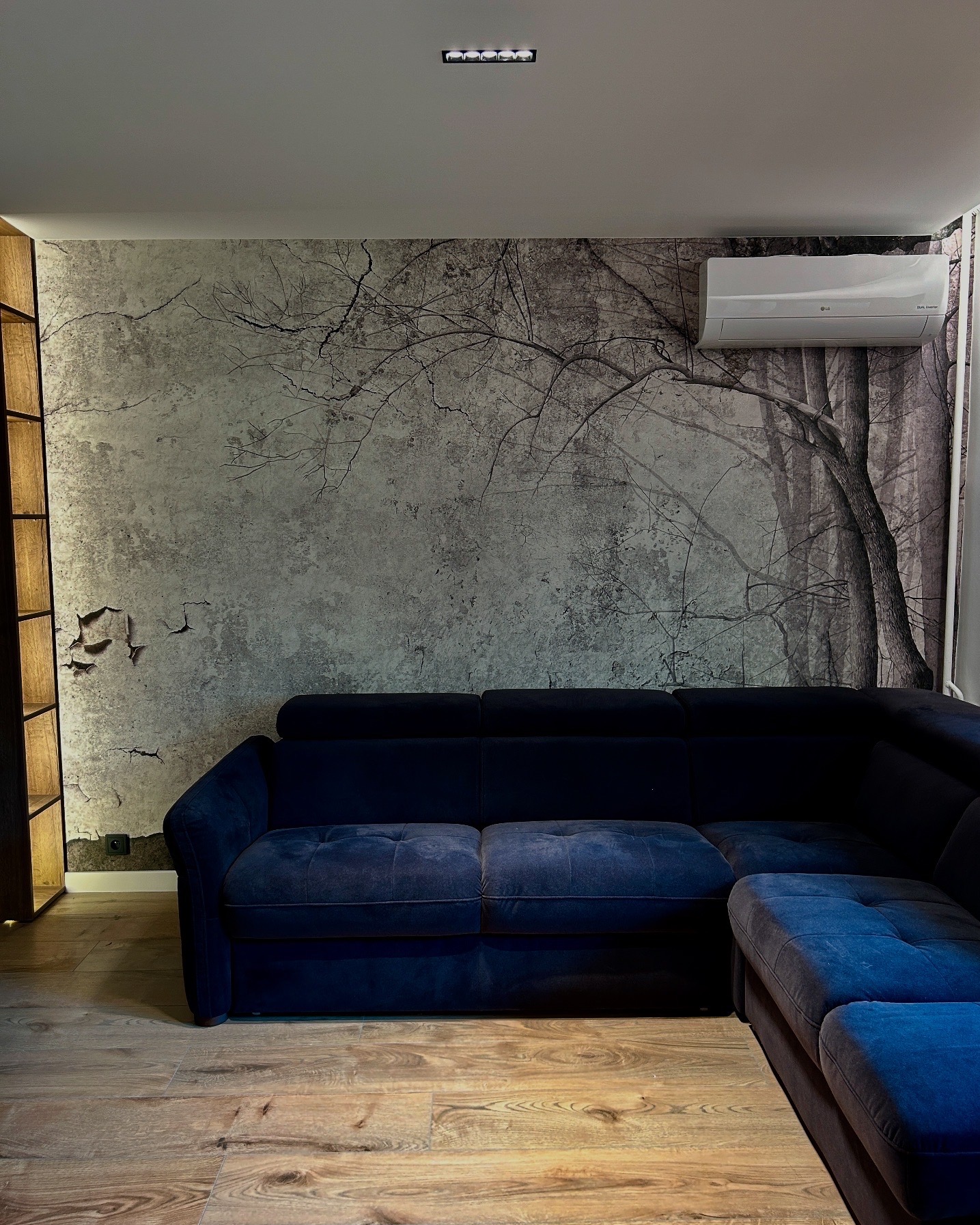 Nowoczesny salon z ciemnoniebieską sofą narożną, drewnianą podłogą i fototapetą imitującą korę drzew oraz klimatyzator na ścianie.