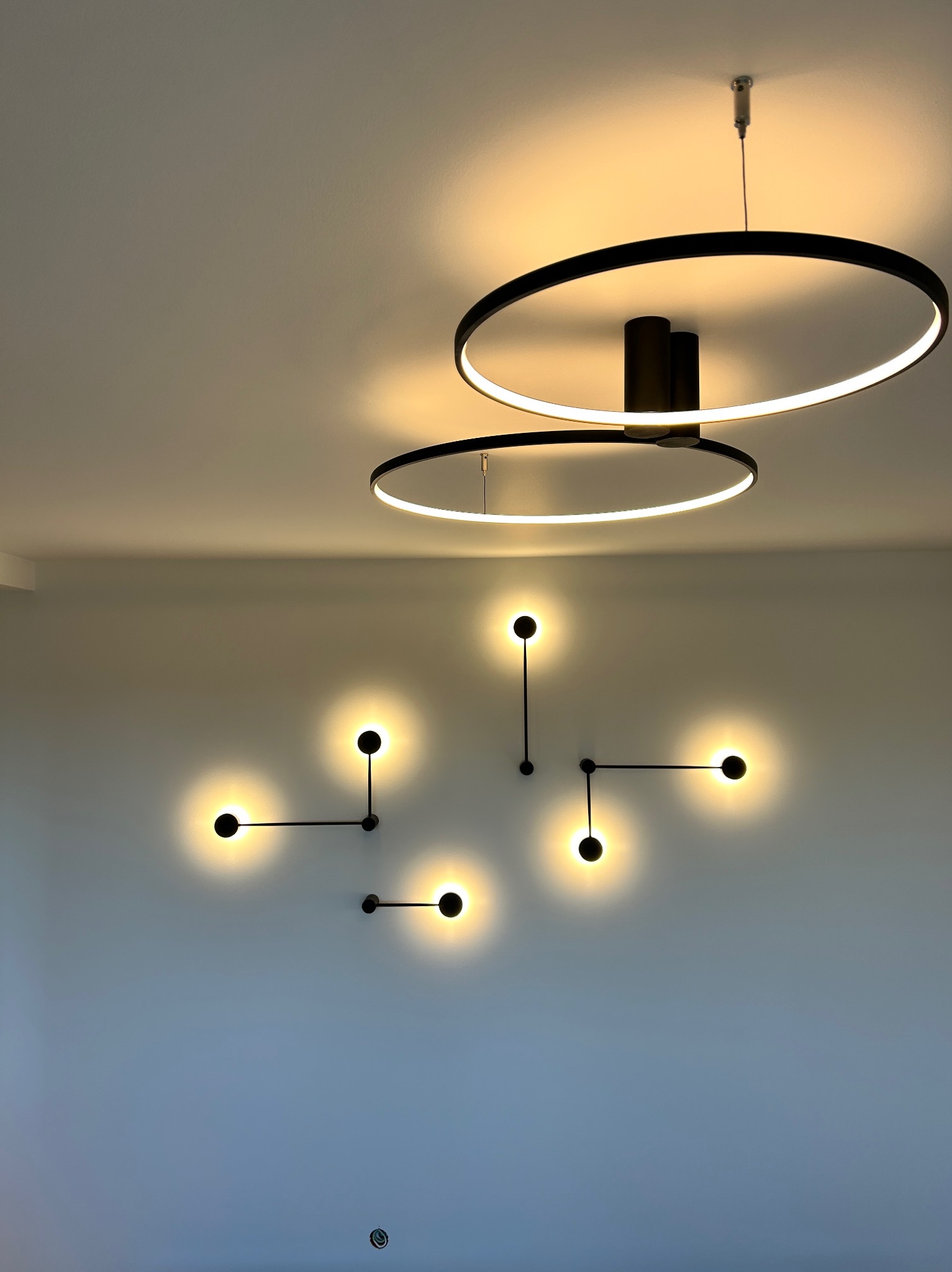 Minimalistyczne oświetlenie wnętrza: dwa okrągłe, wiszące plafony z ledowym światłem oraz kompozycja ściennych lamp punktowych na gładkiej, jasnej ścianie.