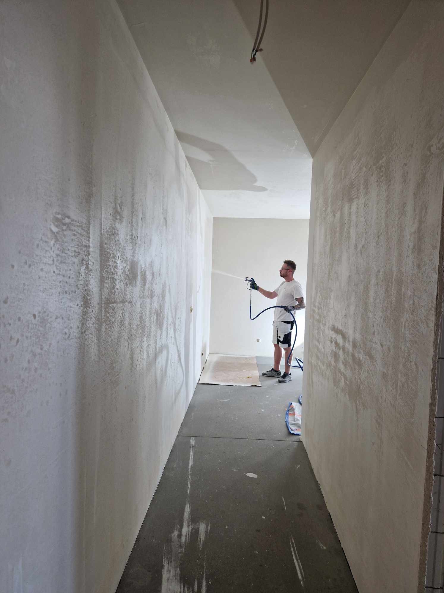 Aplikacja gładzi natryskowej na ścianę w korytarzu, widoczny pracownik z pistoletem natryskowym i ślady świeżej gładzi.