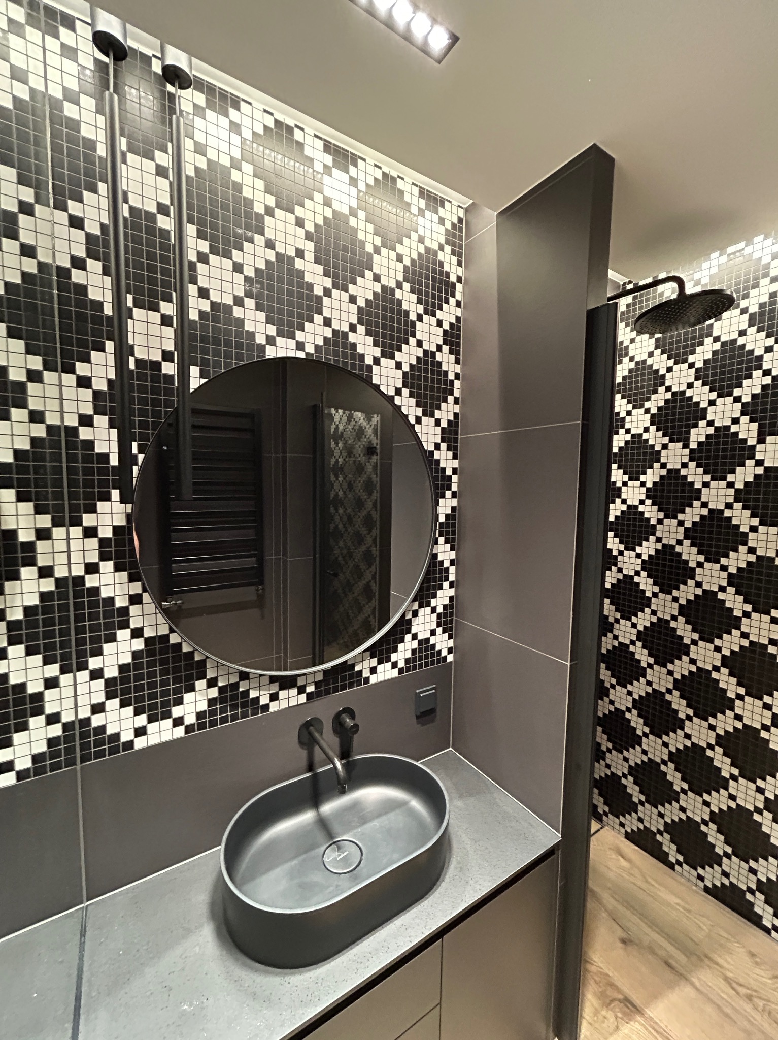 Nowoczesna łazienka w odcieniach szarości i czerni z mozaiką na ścianach, owalną umywalką na blacie i okrągłym lustrem, widoczny fragment kabiny prysznicowej z deszczownicą.