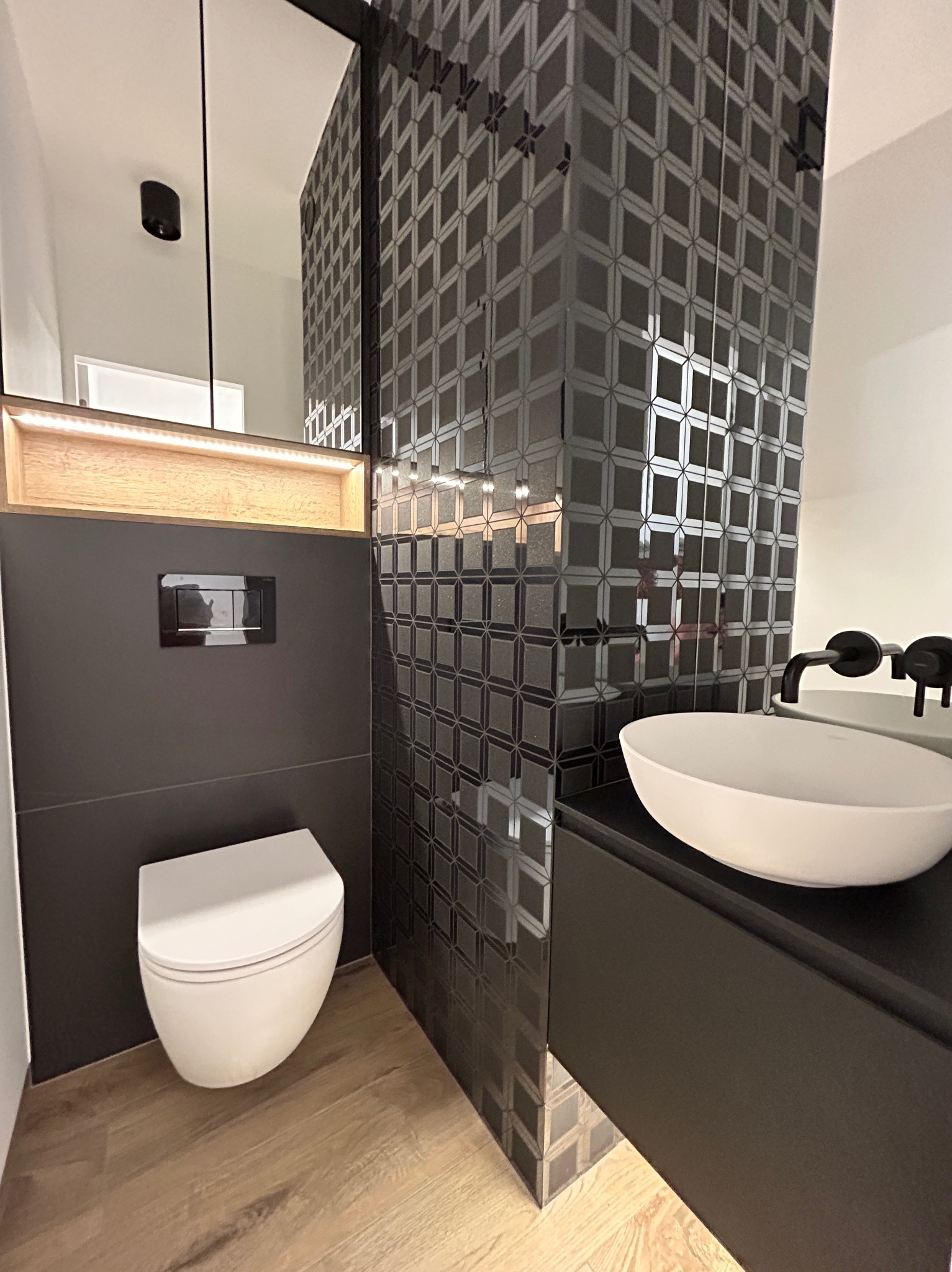 Nowoczesna łazienka z czarnymi, wzorzystymi płytkami na ścianie, białą umywalką nablatową z czarną armaturą, podwieszaną toaletą i drewnopodobną podłogą.