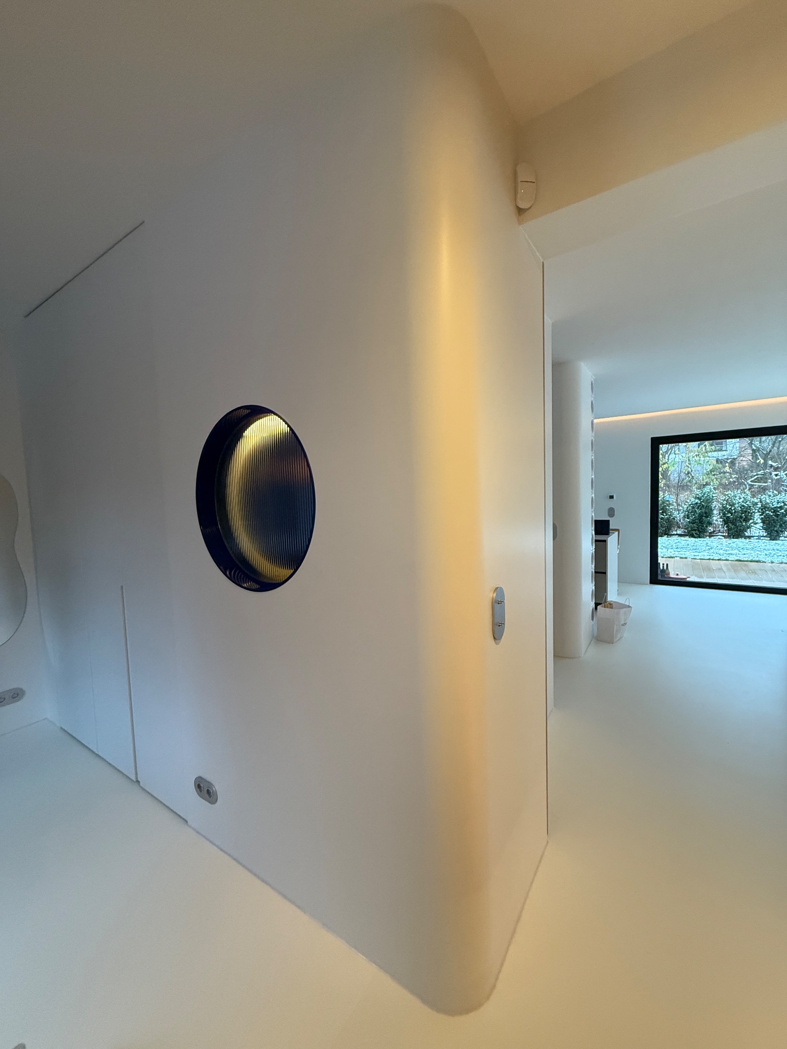 Nowoczesne wnętrze: białe ściany z okrągłym oknem w granatowej ramie, gładka podłoga i widok na ogród przez duże okno. Minimalistyczny design.