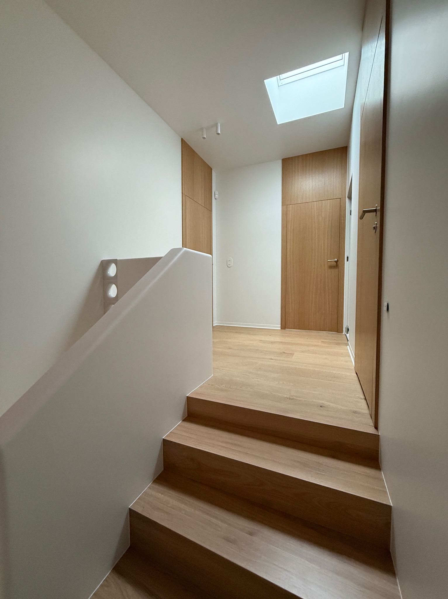 Nowoczesne schody z drewnianymi stopniami i białą balustradą, oświetlone światłem z okna dachowego. Minimalistyczne wnętrze z drewnianymi drzwiami i panelami podłogowymi.