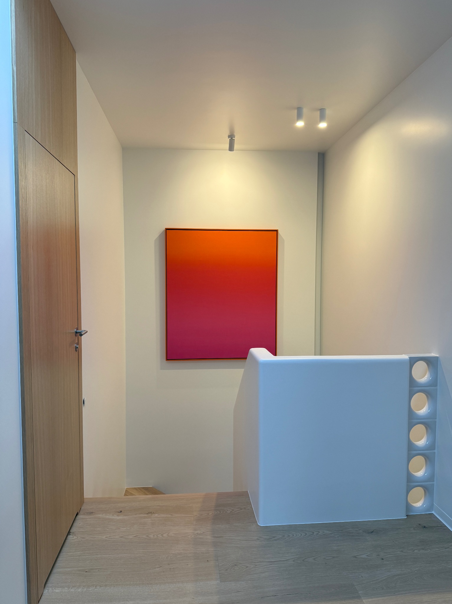 Nowoczesne wnętrze: schody z białą balustradą z otworami, drewniana podłoga i drzwi, ściana z abstrakcyjnym obrazem w odcieniach różu i pomarańczy, oświetlenie punktowe.