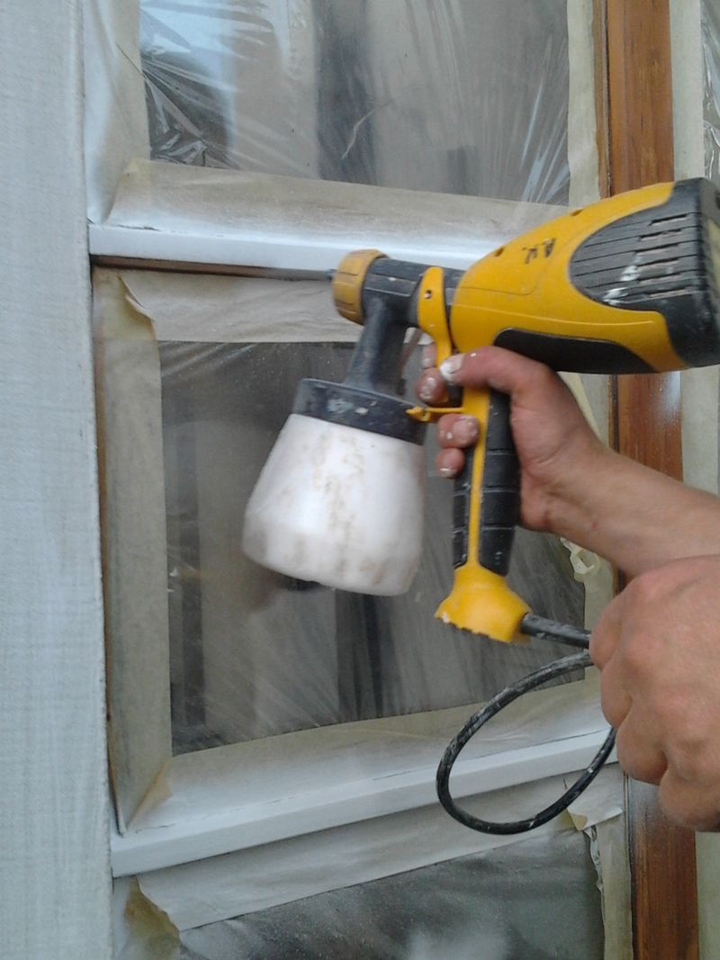 Malowanie okna drewnianego na biało za pomocą pistoletu natryskowego; zabezpieczone folią malarską.