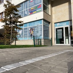 PARTNER TEMPS SPÓŁKA Z OGRANICZONĄ ODPOWIEDZIALNOŚCIĄ - Budynek firmy Partner Temps z widocznymi oknami z reklamami usług, flagami różnych krajów, oraz informacjami kontaktowymi. Przed budynkiem chodnik z kostki brukowej z namalowaną białą linią.