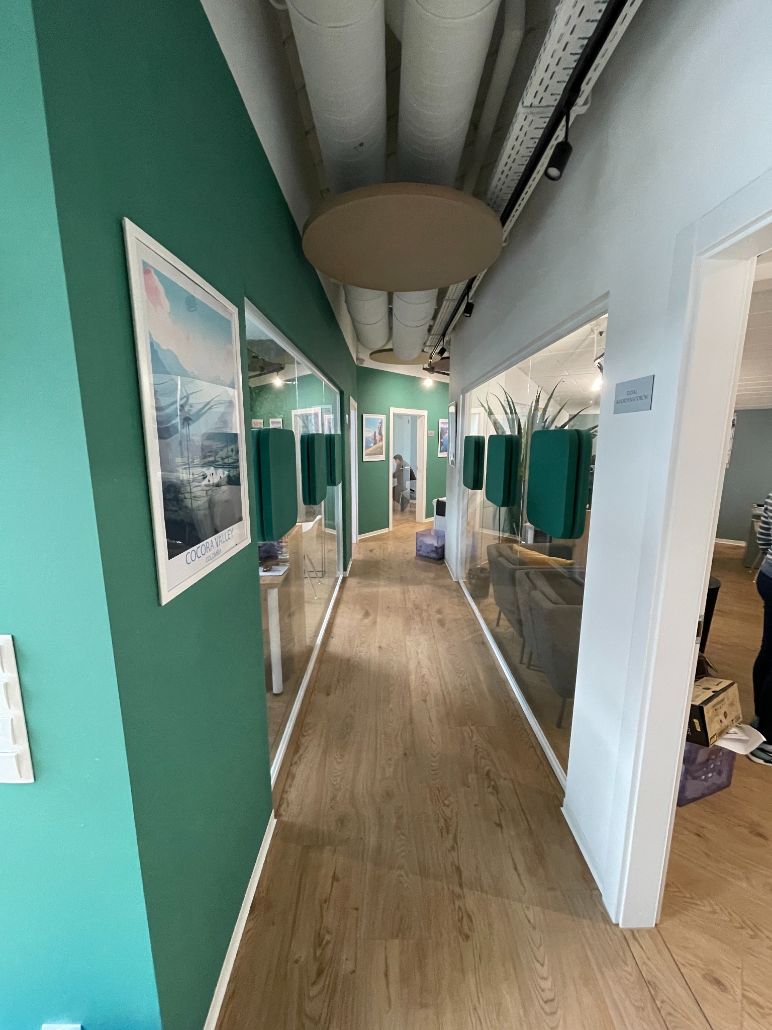 Korytarz biurowy z przeszklonymi ścianami, drewnianą podłogą i zielonymi elementami dekoracyjnymi. Na ścianie wisi plakat z napisem 'Cocora Valley Colombia'.