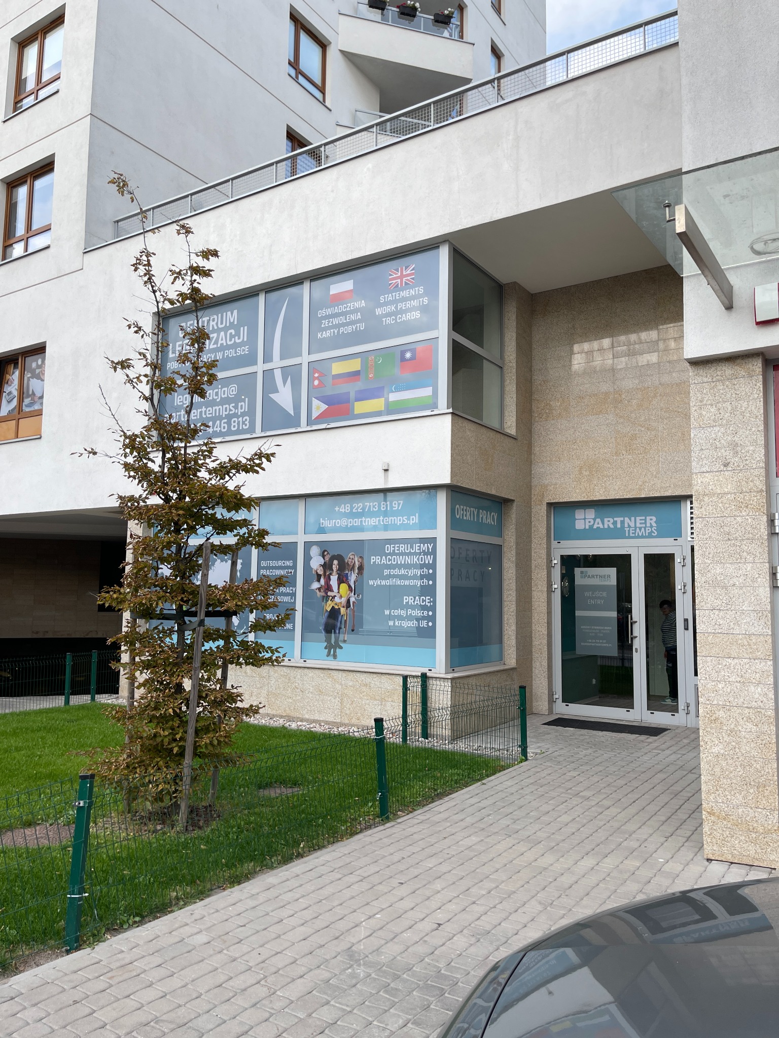 Fasada budynku agencji rekrutacyjnej Partner Temps z widocznymi oknami z ofertami pracy i legalizacji pobytu dla obcokrajowców, drzewko przed wejściem.