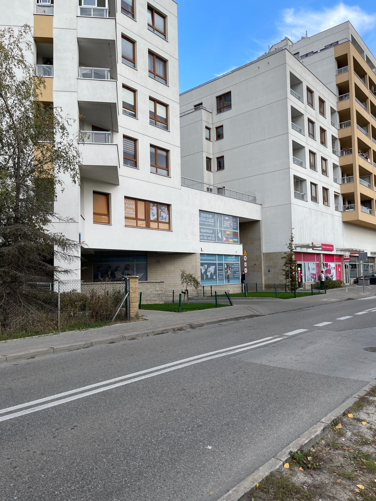 Fasada budynku z biurami usługowych firm, w tym 'Kadry Place' z widocznym logo i danymi kontaktowymi, oraz 'Centrum Legalizacji' z flagami i mapą świata na szybie, widziana z perspektywy ulicy.
