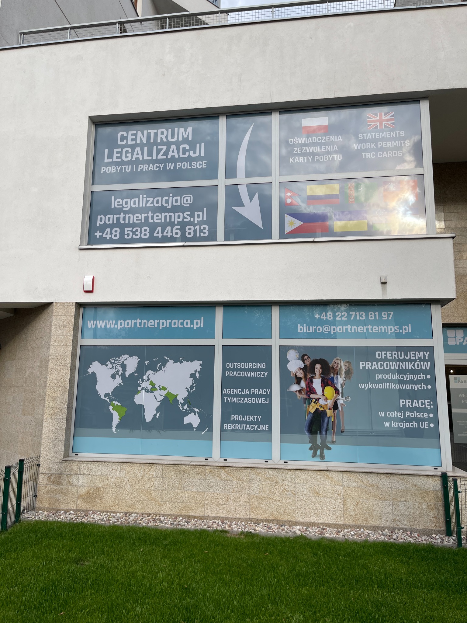 Fasada budynku z reklamami firmy oferującej legalizację pobytu i pracy w Polsce, outsourcing pracowniczy oraz usługi agencji pracy tymczasowej. Na oknach widoczne mapy świata i zdjęcia pracowników...
